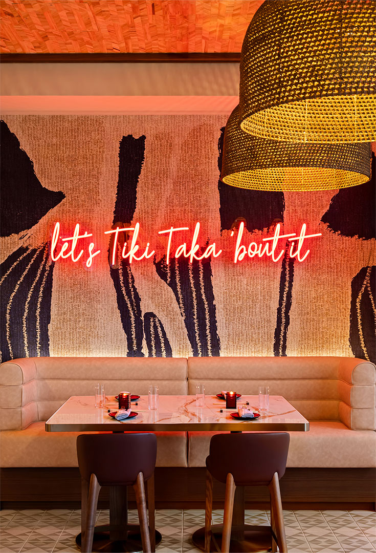 Neon sign reading “let’s Tiki Taka ’bout it” above a cozy booth with modern wall art and ambient lighting.