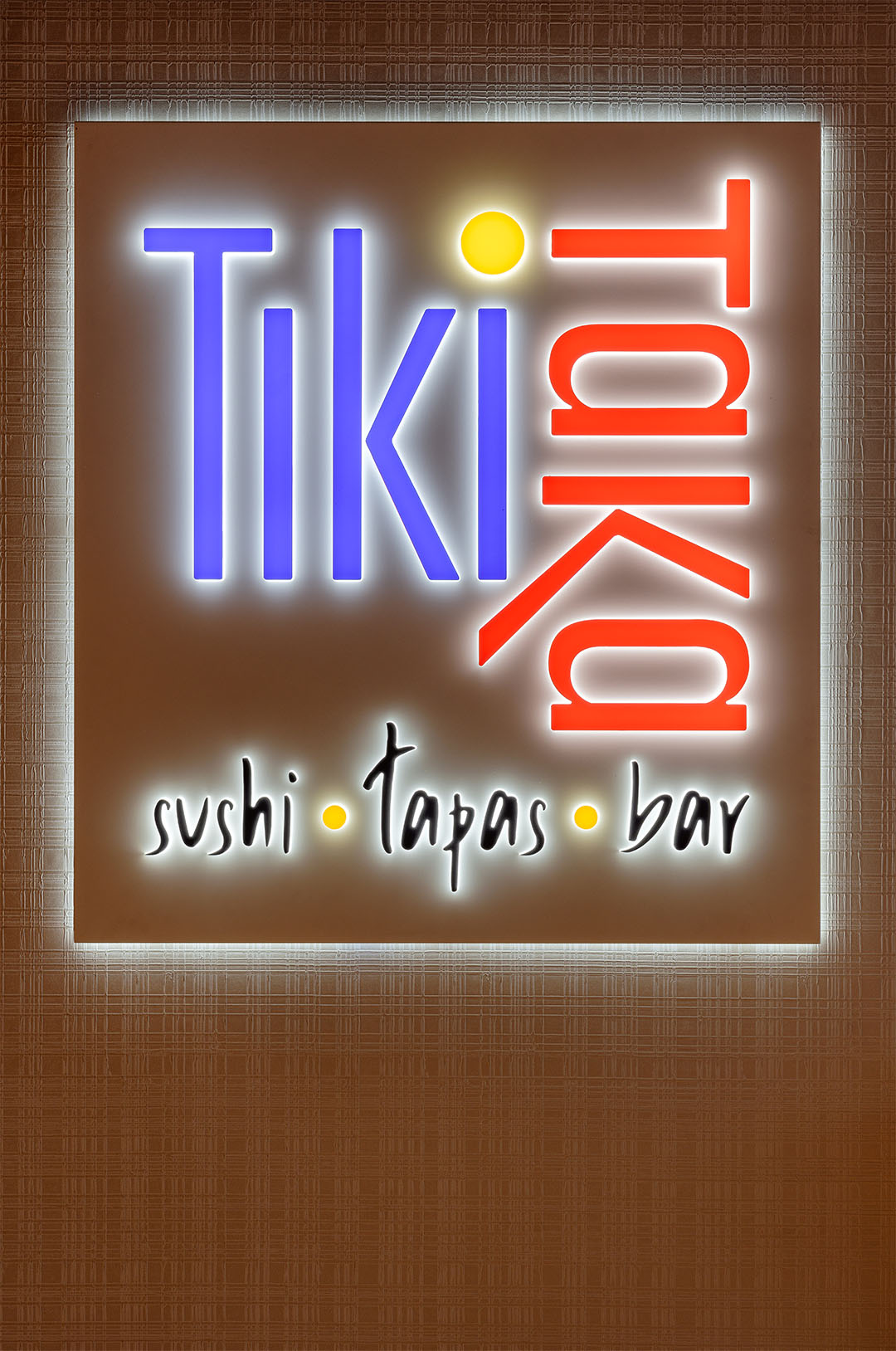 Illuminated Tiki Taka logo with playful typography and the tagline “sushi · tapas · bar” on a textured wall.